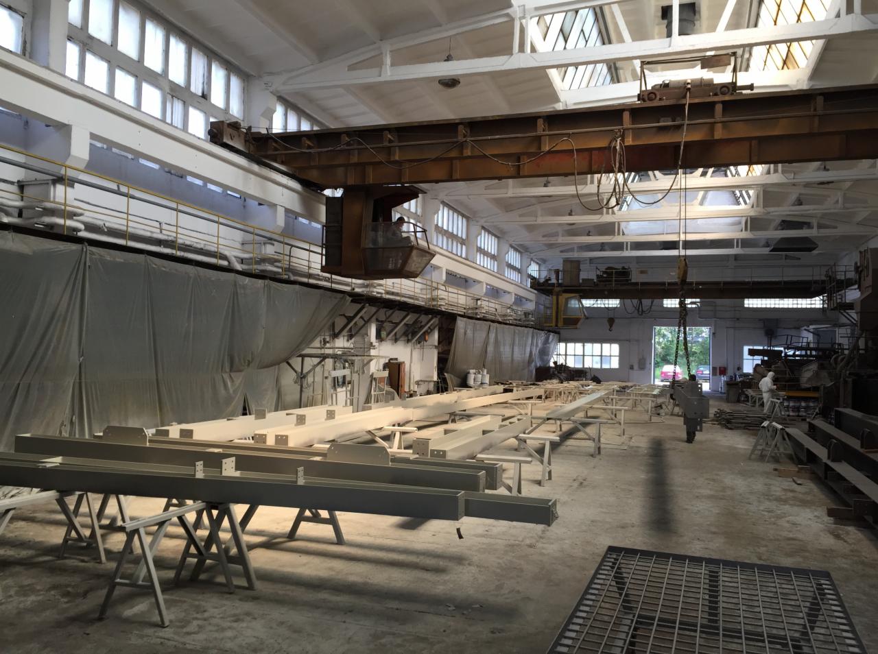 Wnętrze hali produkcyjnej z suwnicą i pomalowanymi na szaro metalowymi elementami konstrukcyjnymi ustawionymi na kozłach.