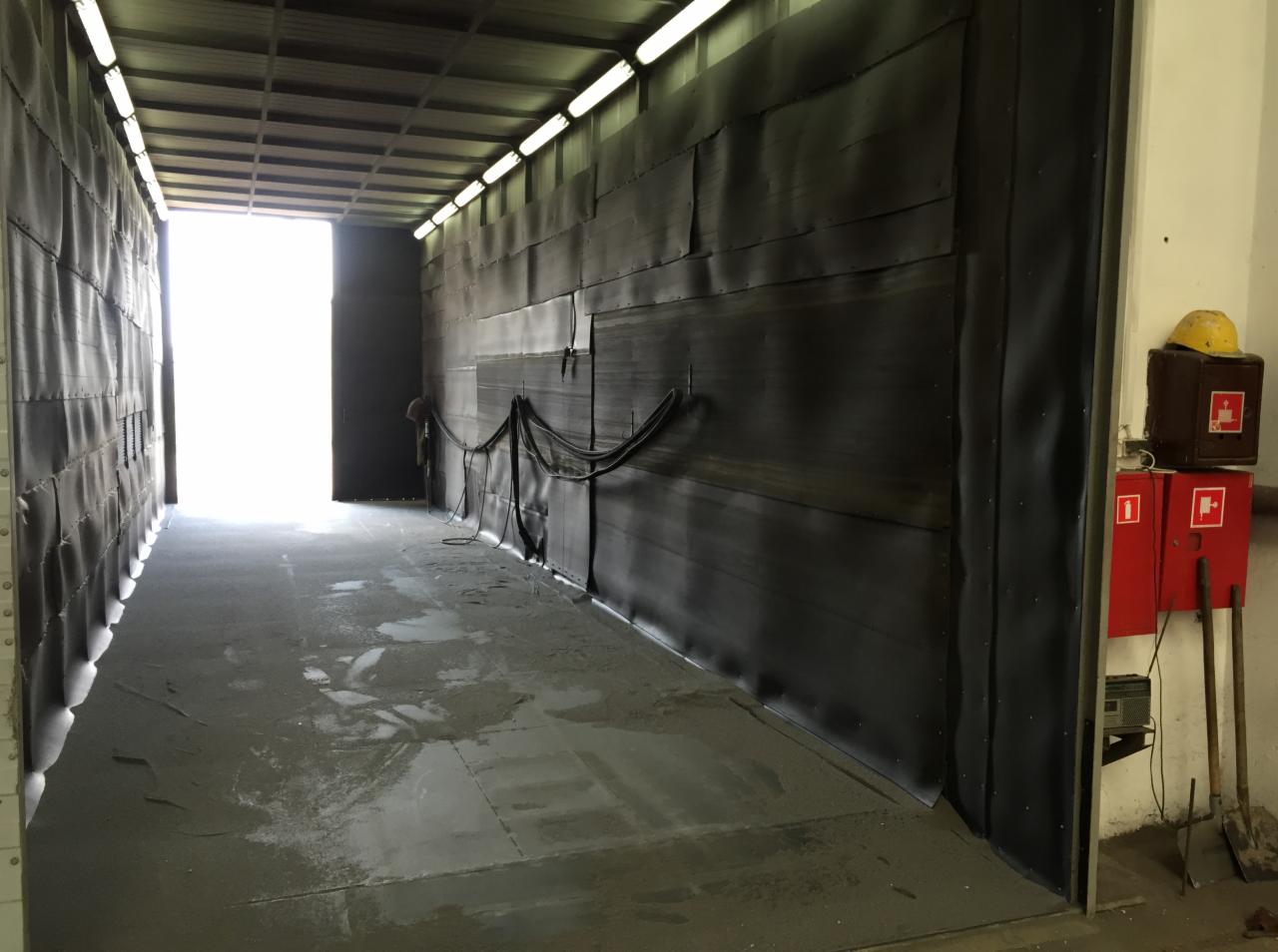Wnętrze hali z ciemnymi ścianami, prawdopodobnie przygotowanej do piaskowania lub malowania, widoczne przewody i sprzęt BHP przy ścianie.