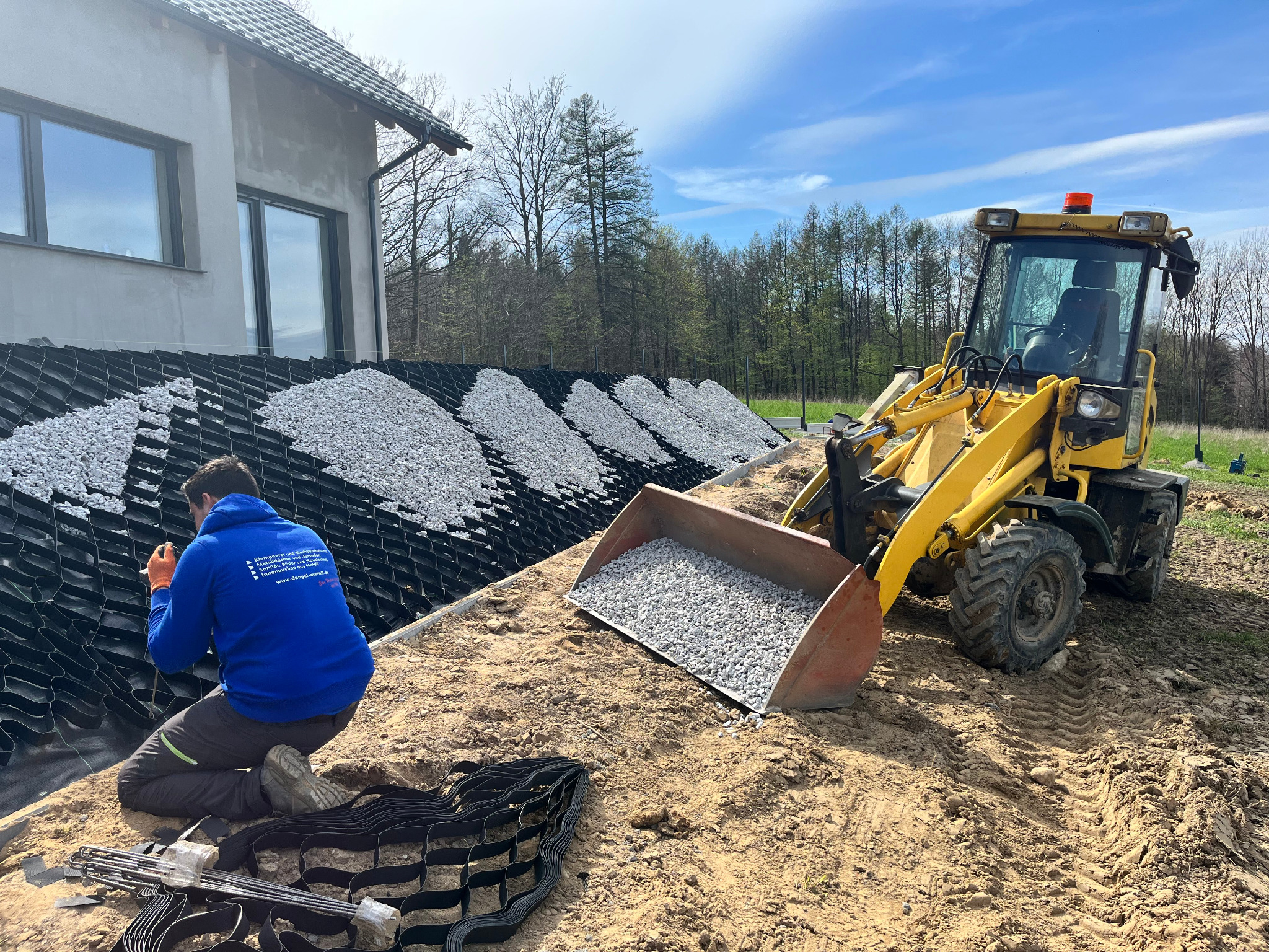 Pracownik montuje geokratę na zboczu, obok stoi żółta ładowarka z gruzem. W tle dom w stanie surowym i drzewa. Prace ziemne w słoneczny dzień.