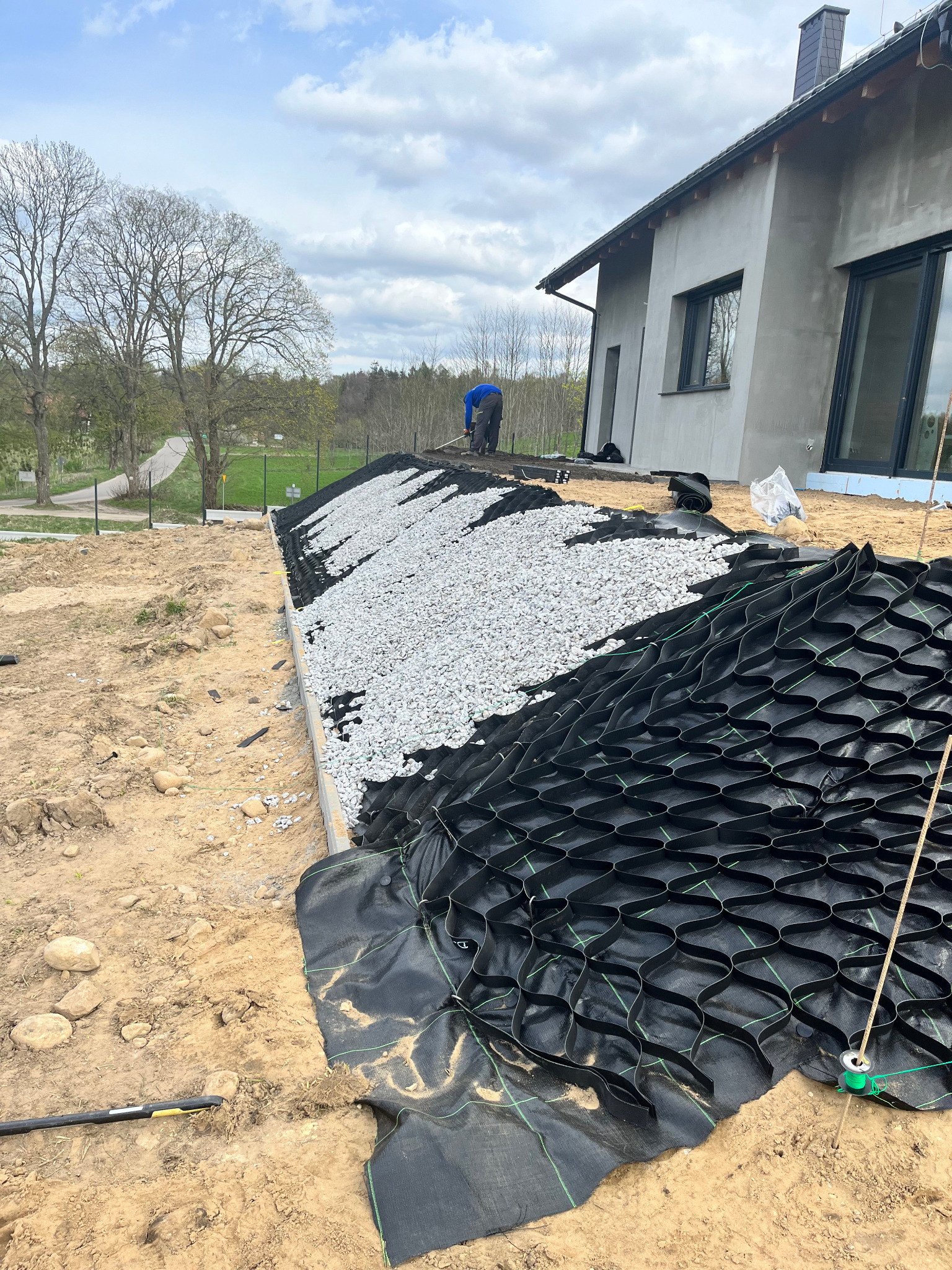Układanie geokraty i kamienia na przygotowanym terenie przy domu. Widoczny pracownik w oddali. Etap prac ziemnych przed zagospodarowaniem ogrodu.