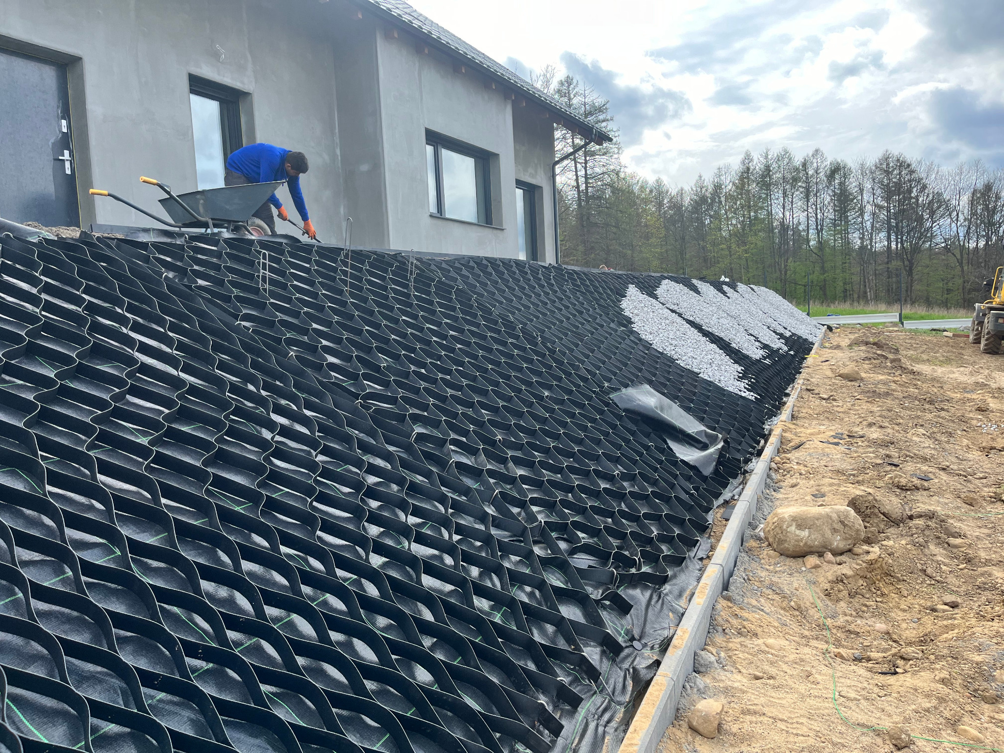 Geosyntetyczna siatka na zboczu wzmacnia grunt pod domem. Pracownik z taczką i narzędziem. Częściowo wypełniona białym żwirem. Stabilizacja gruntu.