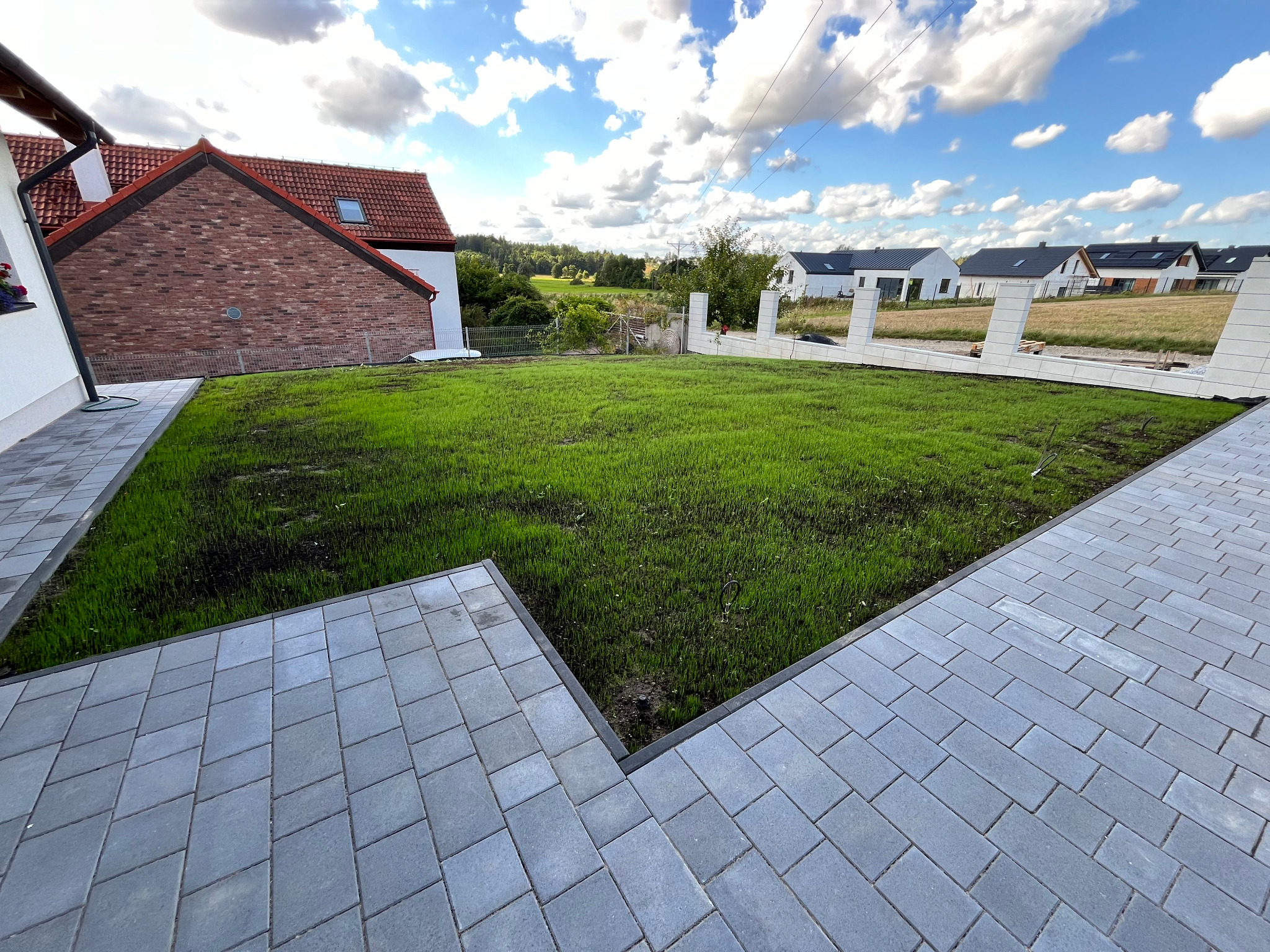 Świeżo założony trawnik z młodą trawą, otoczony szarą kostką brukową, na tle domu z czerwoną dachówką i błękitnego nieba z chmurami.