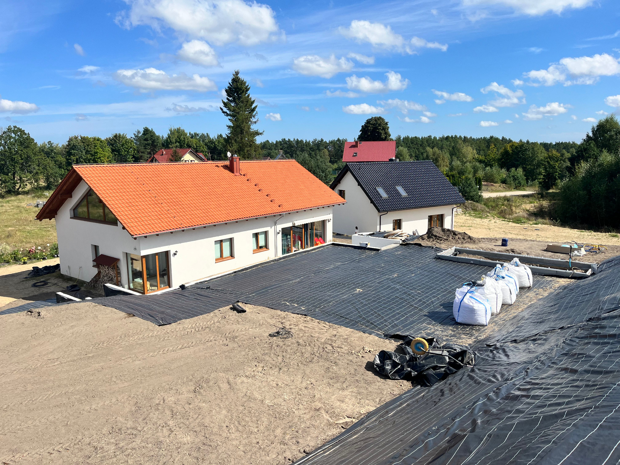 Przygotowanie terenu pod trawnik: rozłożona czarna mata, białe worki z ziemią, dwa domy w tle pod błękitnym niebem. Widok z góry na plac budowy.