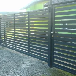 Nowoczesne ogrodzenie metalowe z poziomymi panelami w kolorze grafitowym, brama wjazdowa z klamką, podłoże żwirowe, w tle dom z zieloną elewacją.