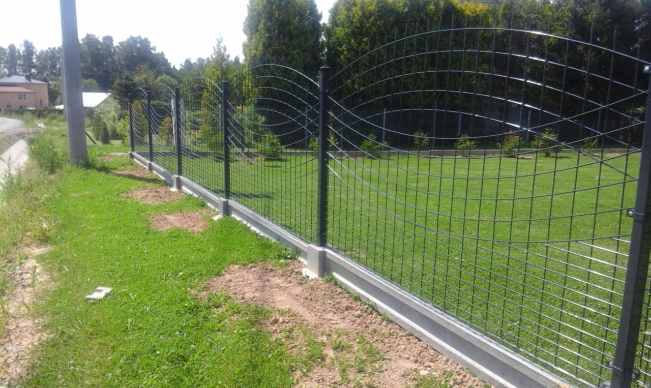 Nowoczesne ogrodzenie z siatki z ozdobnym łukiem, osadzone na betonowej podmurówce, widok z boku na tle zielonego trawnika i drzew