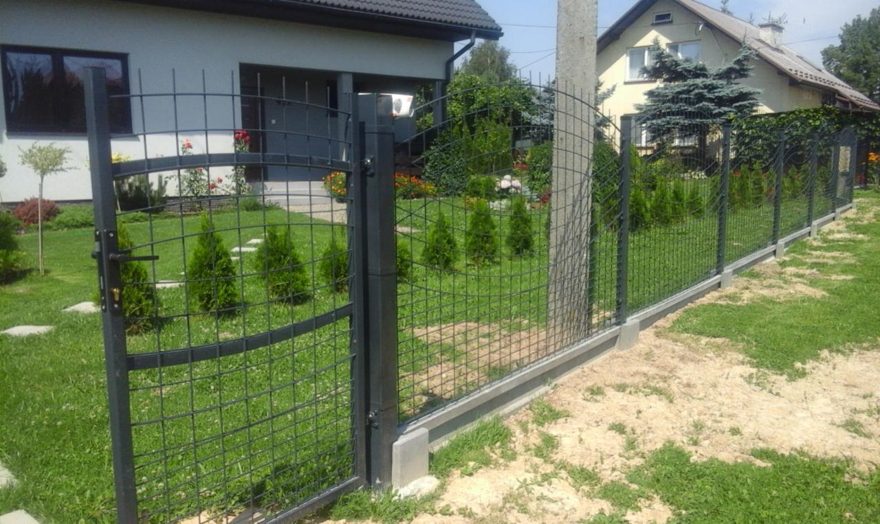 Nowoczesne ogrodzenie z siatki panelowej w kolorze grafitowym, z furtką i podmurówką betonową, otoczone zielonym trawnikiem i tujami, na tle domu jednorodzinnego.