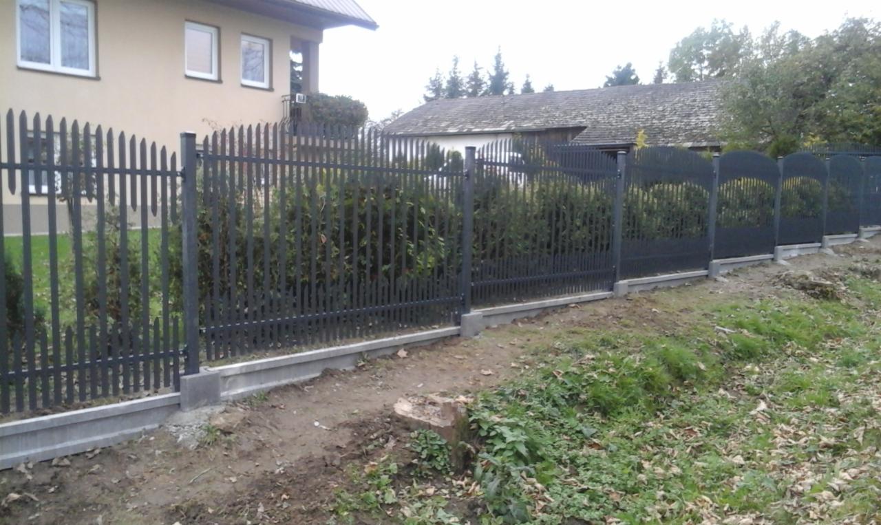 Ciemnoszary płot z metalowych prętów na betonowej podmurówce, z roślinnością w tle i fragmentami budynku mieszkalnego w oddali.