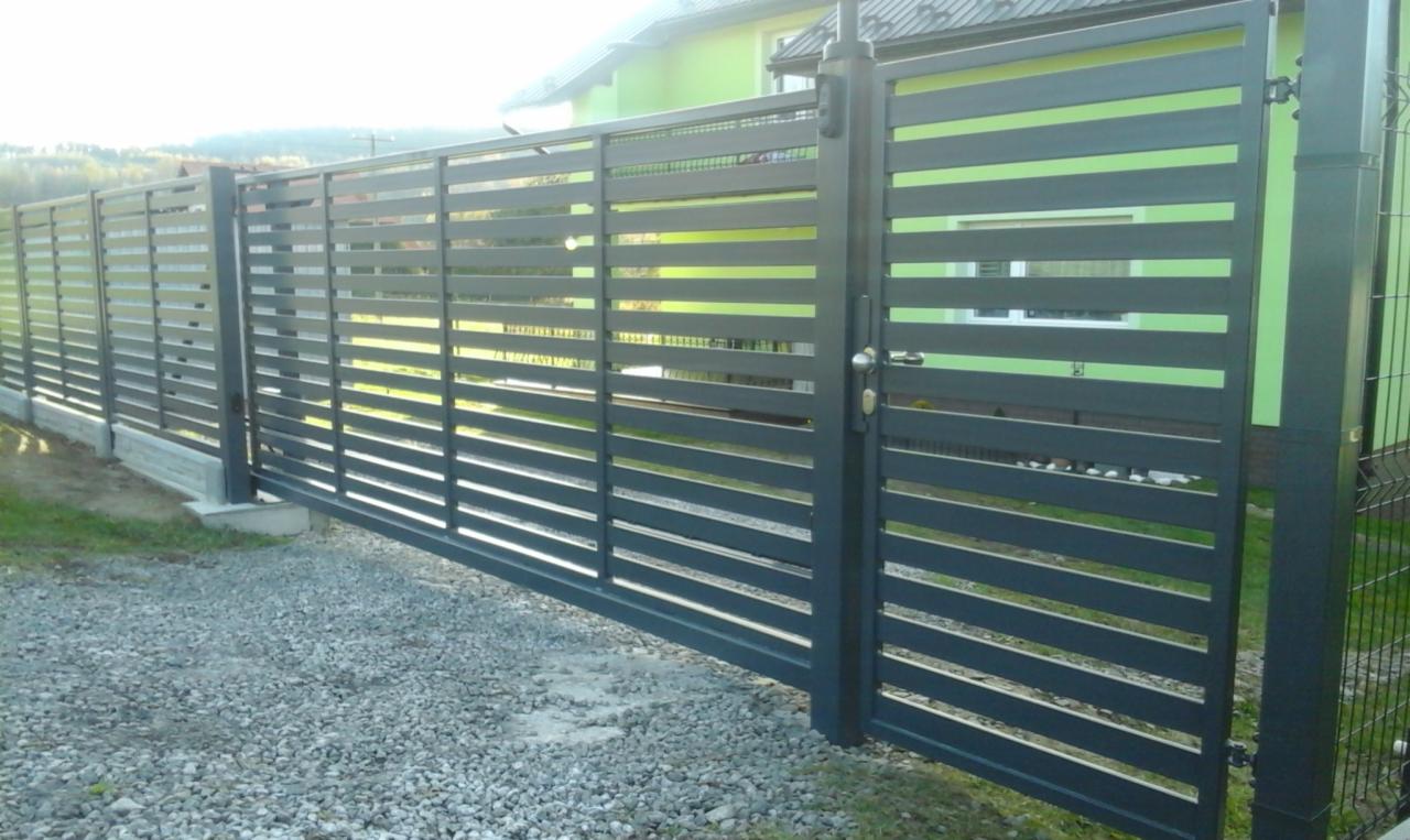 Nowoczesne ogrodzenie metalowe z poziomymi panelami w kolorze grafitowym, brama wjazdowa z klamką, podłoże żwirowe, w tle dom z zieloną elewacją.