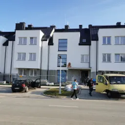 Nowoczesny budynek wielorodzinny w trakcie wykończenia elewacji, z widocznymi pracownikami, materiałami budowlanymi, samochodem osobowym i dostawczym na placu budowy.