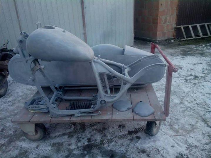 Rama motocykla z bocznym wózkiem, pokryta szarym podkładem, umieszczona na wózku transportowym. Widoczne elementy zawieszenia i zbiornik paliwa. Tło stanowi blaszany garaż i ceglany budynek.