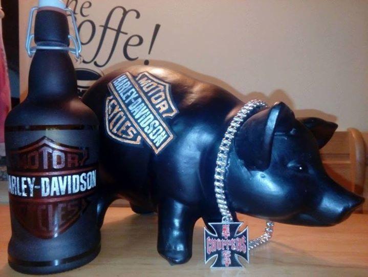 Czarny ceramiczny prosiak z logo Harley-Davidson i srebrnym łańcuchem z krzyżem Choppers, obok butelka z logo Harley-Davidson na drewnianym blacie.