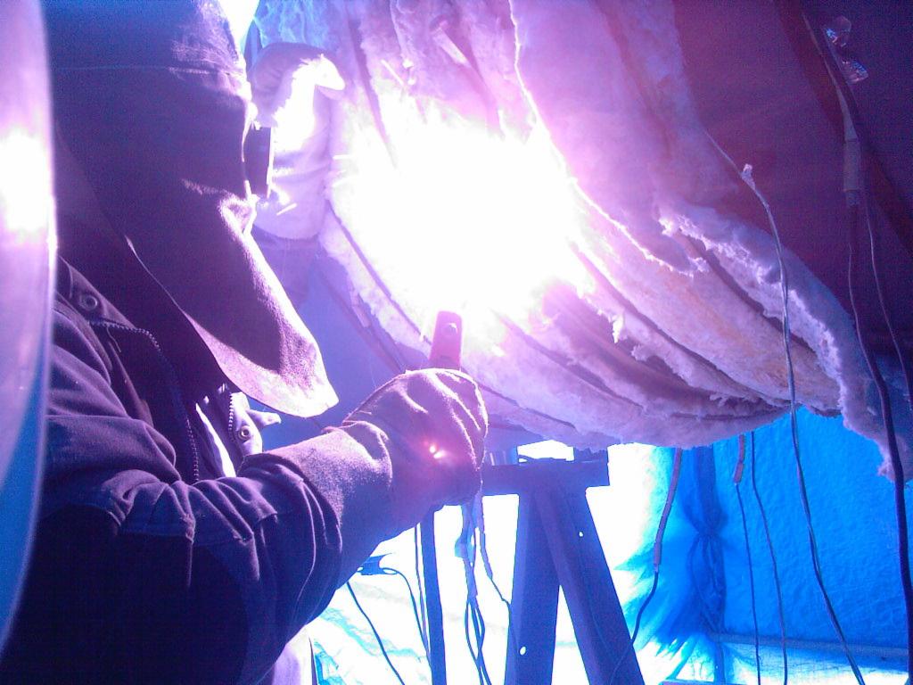 Pracownik w masce spawalniczej i rękawicach wykonuje spawanie konstrukcji z izolacją na rusztowaniu, widoczne okablowanie i niebieska folia ochronna.