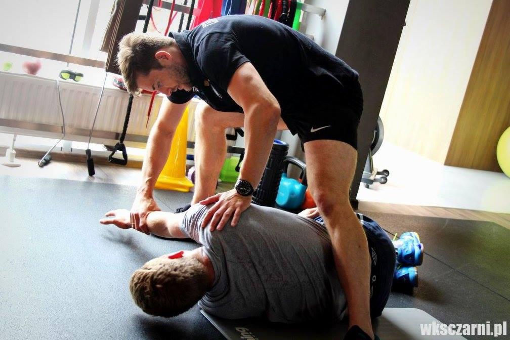 Mężczyzna w czarnych spodenkach i koszulce uciska ramię leżącego na macie mężczyzny, w tle sprzęt do ćwiczeń i żółty pachołek.