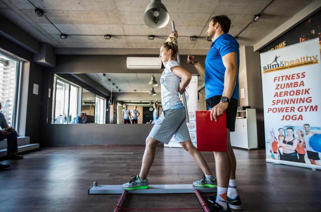 Kobieta w sportowym stroju wykonuje ćwiczenie postawy z pomocą trenera i specjalistycznego sprzętu w jasnym wnętrzu studia fitness, widoczny plakat z ofertą zajęć.