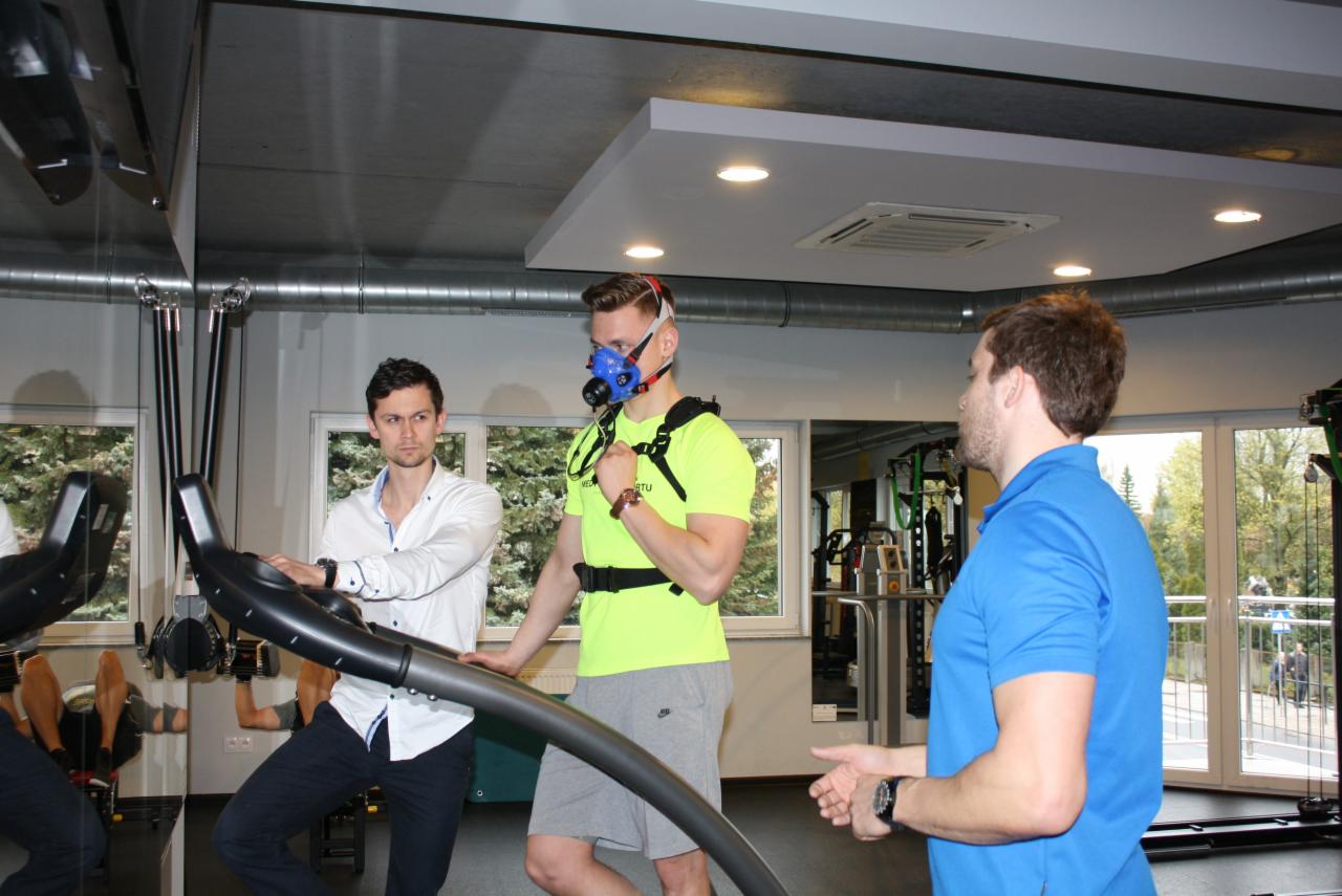 Mężczyzna z maską tlenową na twarzy, w neonowej koszulce, na bieżni, monitorowany przez dwóch mężczyzn w jasnych koszulach w siłowni.