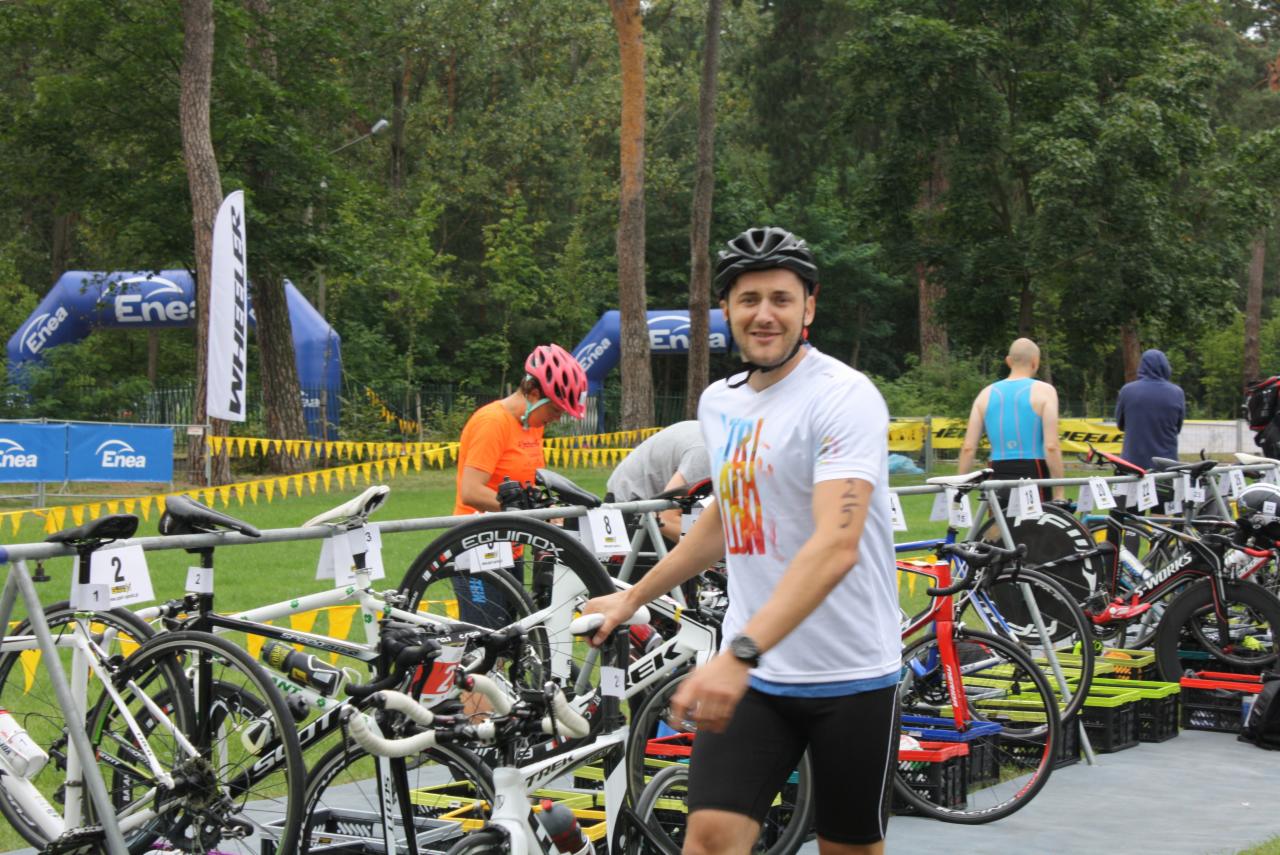 Uśmiechnięty mężczyzna w kasku rowerowym i koszulce triathlonowej stoi przy rzędzie rowerów na stanowisku dla zawodników, w tle widoczne banery reklamowe i osoby przygotowujące się do zawodów.