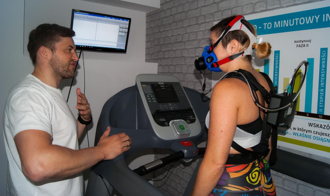 Kobieta z maską pomiarową na twarzy, podłączona do aparatury na plecach, biegnie na bieżni, monitorowana przez mężczyznę w białej koszulce, który obserwuje wyniki na ekranie komputera. W tle ściana...