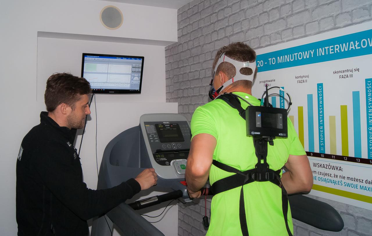 Mężczyzna w masce tlenowej i z urządzeniem pomiarowym na plecach ćwiczy na bieżni pod nadzorem, na ścianie wykres interwałów treningowych i monitor z wynikami pomiarów.