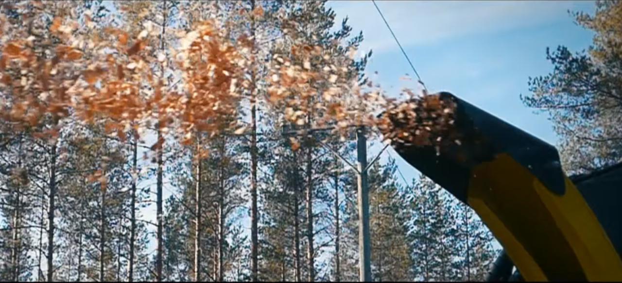 Zimowe odśnieżanie terenu przy użyciu dmuchawy wyrzucającej śnieg na tle lasu sosnowego i błękitnego nieba.