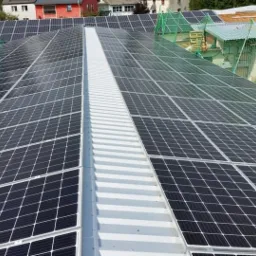 Rozległa instalacja fotowoltaiczna na dachu budynku, widok z góry, z widocznymi panelami i elementami konstrukcyjnymi oraz siatką ochronną w tle.