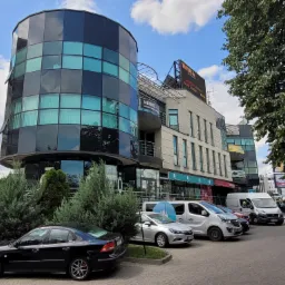 Nowoczesny budynek biurowy z cylindryczną przeszkloną częścią, zaparkowane samochody na pierwszym planie, reklama 'Taurus' na dachu.