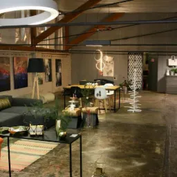 Przestronne wnętrze biura z szarą sofą, pomarańczowym fotelem, drewnianymi stołami, designerskim oświetleniem w postaci okręgów i wiszących lamp oraz betonową podłogą, ukazujące aranżację strefy...