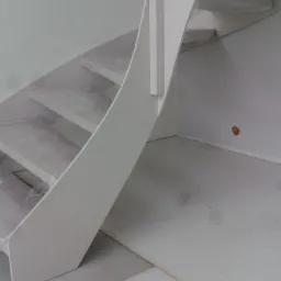 Białe, minimalistyczne schody z zabiegiem, zabezpieczone folią ochronną podczas montażu, widoczne na tle surowego wnętrza w trakcie wykończenia.