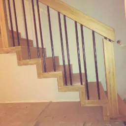 Nowoczesne schody z jasnego drewna z czarnymi, metalowymi balustradami, widok z dołu, w tle jasna ściana.