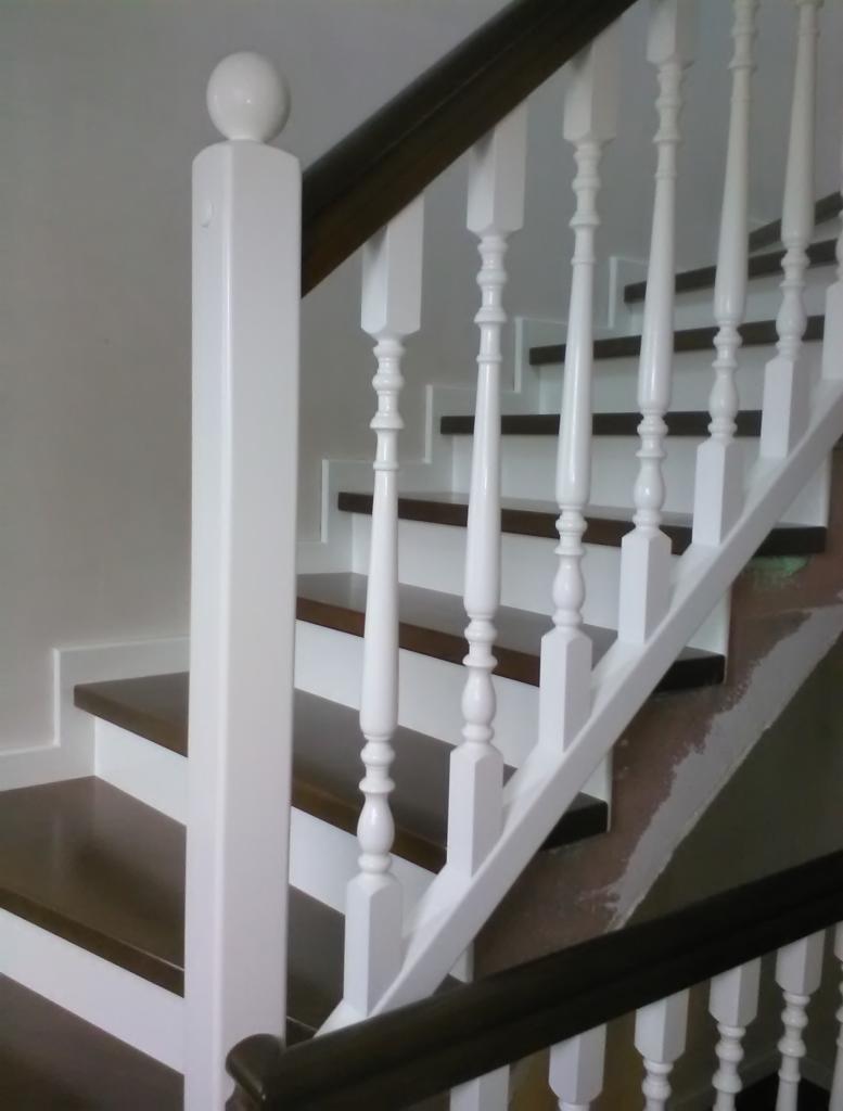 Kręte schody z ciemnobrązowymi stopniami i białymi podstopnicami, balustradą z białych tralek i ciemnobrązową poręczą, zakończone białym słupkiem z kulą.