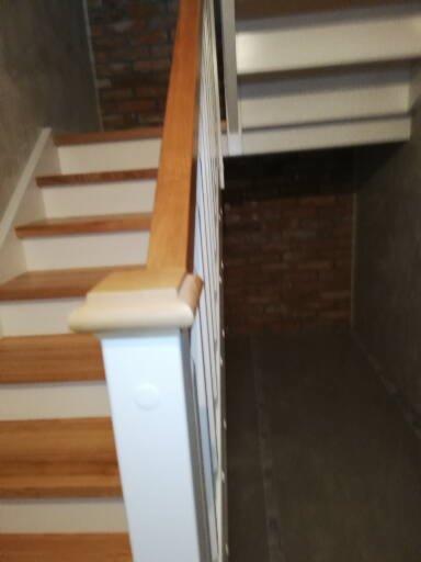 Fragment drewnianych schodów z białą balustradą i jasnobrązową poręczą, widok z dołu, ściana z cegły w tle.