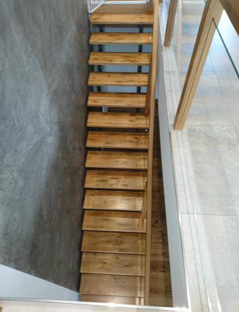 Wąskie, ażurowe schody z jasnego drewna, z minimalistyczną, szklaną balustradą i szarą ścianą w tle, widziane z góry.