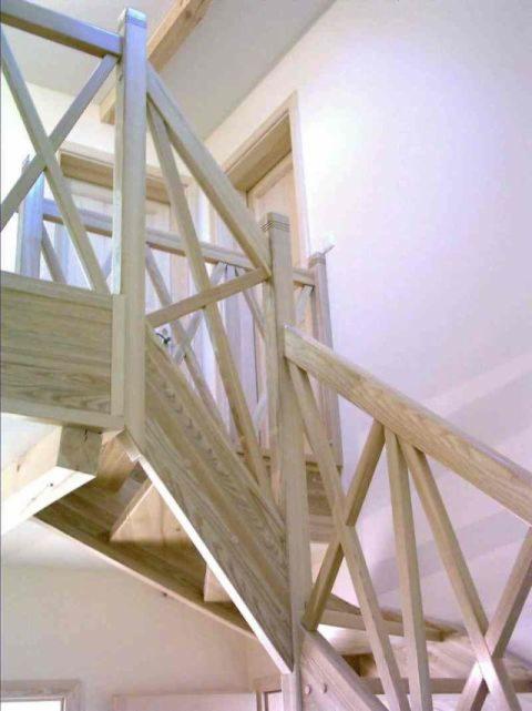 Jasne, drewniane schody z balustradą w kształcie krzyży, widok od dołu, w tle biała ściana i drzwi.