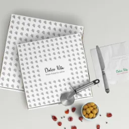 Dwie białe pudełka na pizzę z czarnym wzorem trójkątów i napisem 'Dolce Vita', serwetka z logo, nóż do pizzy, miseczka z oliwkami i suszone pomidory na jasnym tle.
