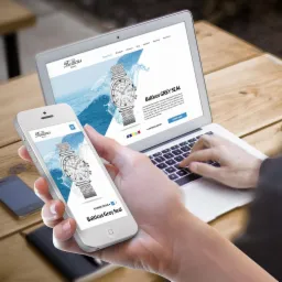 Laptop i smartfon prezentują responsywny wygląd strony internetowej Balticus Watches z zegarkiem Grey Seal na drewnianym stole, dłoń pisze na klawiaturze laptopa.