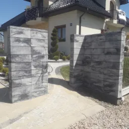 Nowoczesne wejście do posesji z szarych, betonowych element&oacute;w, z widoczną kostką brukową na podjeździe i fragmentem ogrodzenia panelowego z siatki.