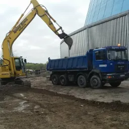 Żółta koparka Komatsu ładuje ziemię do niebieskiej ciężarówki MAN na placu budowy, w tle widoczny budynek o metalowej elewacji.