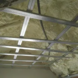 Poddasze z metalową konstrukcją sufitu podwieszanego, wypełnione jasną, nierówną warstwą izolacji natryskowej.