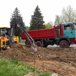 Żółta koparko-ładowarka Volvo BL71B nabiera łyżką ziemię, którą obok ładuje czerwona ciężarówka MAN E7/F2000 na tle zieleni drzew i wiosennych kwiatów.
