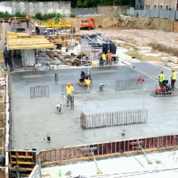 Wylewanie i wygładzanie betonu na budowie, widoczne zbrojenia, ekipa w odblaskowych kamizelkach, betoniarka i koparka w tle.