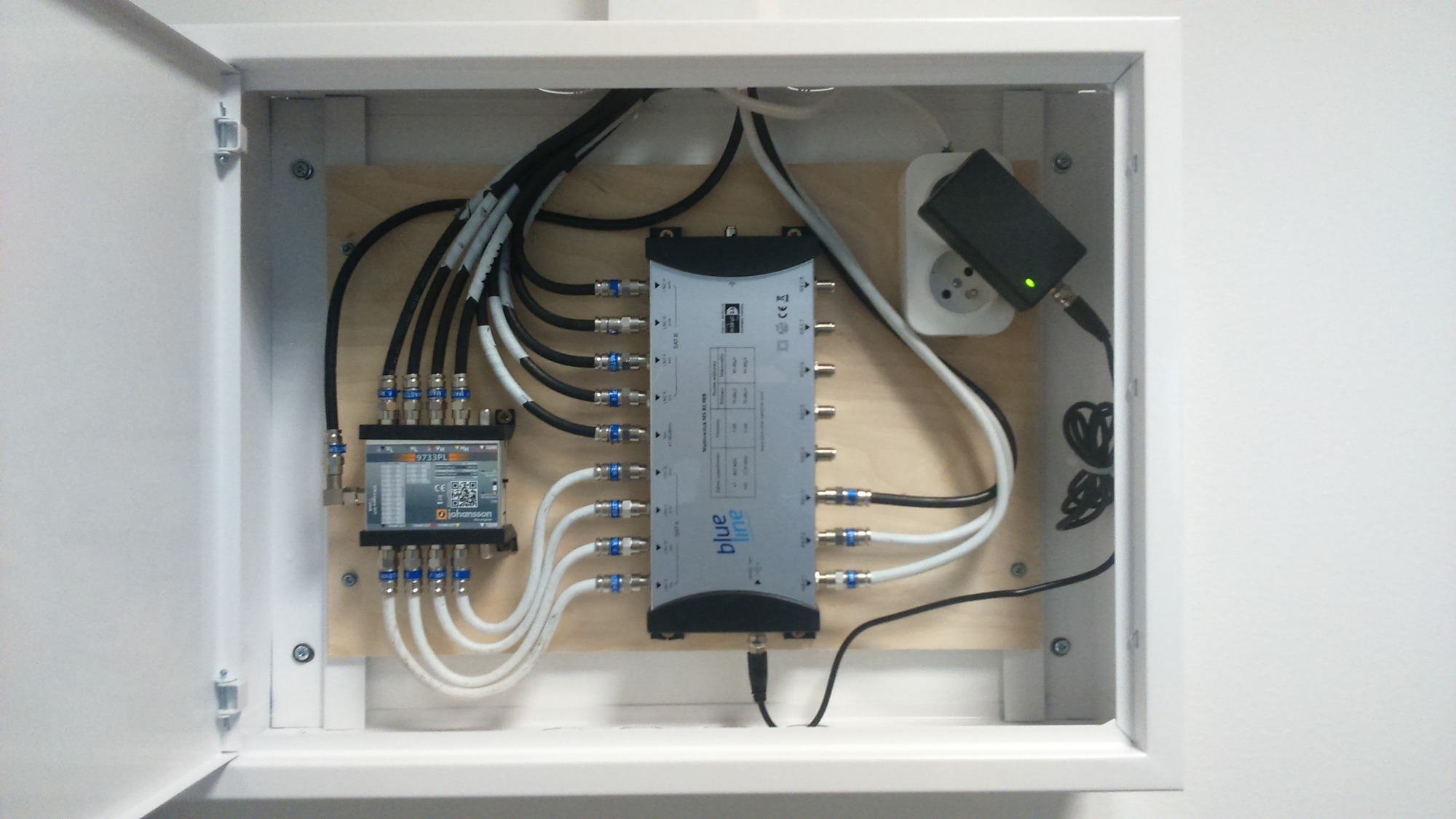 Rozdzielacz antenowy Blue Line w białej szafce, z podłączonymi kablami koncentrycznymi i zasilaczem sieciowym. Widoczny również mniejszy rozdzielacz Johansson.