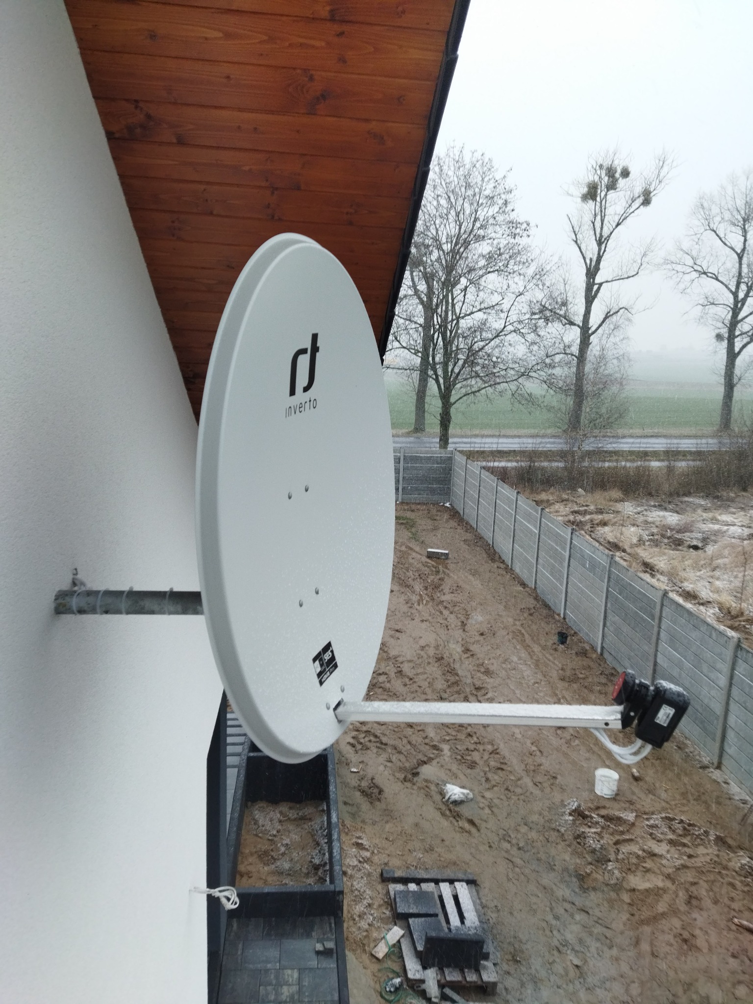 Biała antena satelitarna marki Inverto zamontowana na ścianie domu z widokiem na pochmurny krajobraz i betonowy płot.