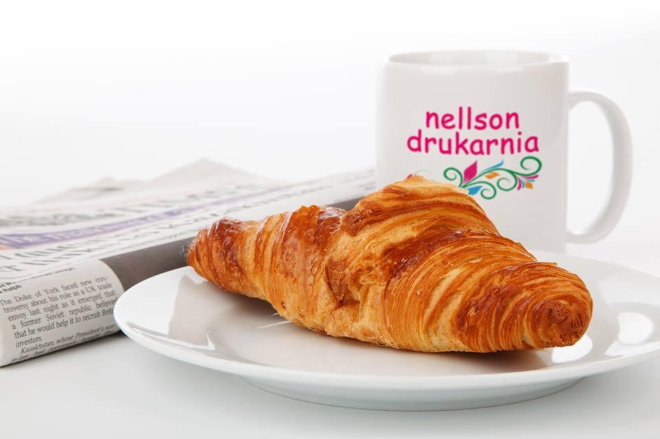 Śniadanie z rogalikiem na talerzu, poranna gazeta i kubek z kolorowym logo drukarni 'nellson drukarnia' na białym tle.