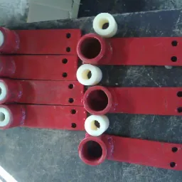 Czerwone, metalowe elementy z przyspawanymi uchwytami i włożonymi białymi tulejami z tworzywa sztucznego, ułożone rzędami na brudnym stole warsztatowym.