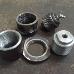 PPUH WOMAR - Zestaw metalowych elementów z gwintami, w tym tuleje, pierścień i element z kulką, leżących na szorstkiej, metalowej powierzchni.