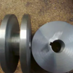 Dwa metalowe koła pasowe o różnych rozmiarach, jedno z otworem na wpust, leżące na brudnej powierzchni warsztatowej.