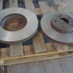PPUH WOMAR - Dwa metalowe dyski hamulcowe, jeden z piastą z otworami na śruby, leżące na drewnianej palecie w warsztacie.