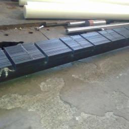 PPUH WOMAR - Czarny, stalowy profil z wyfrezowanymi rowkami, leżący na betonowej podłodze w warsztacie, z widocznymi białymi wałkami z tworzywa sztucznego w tle.