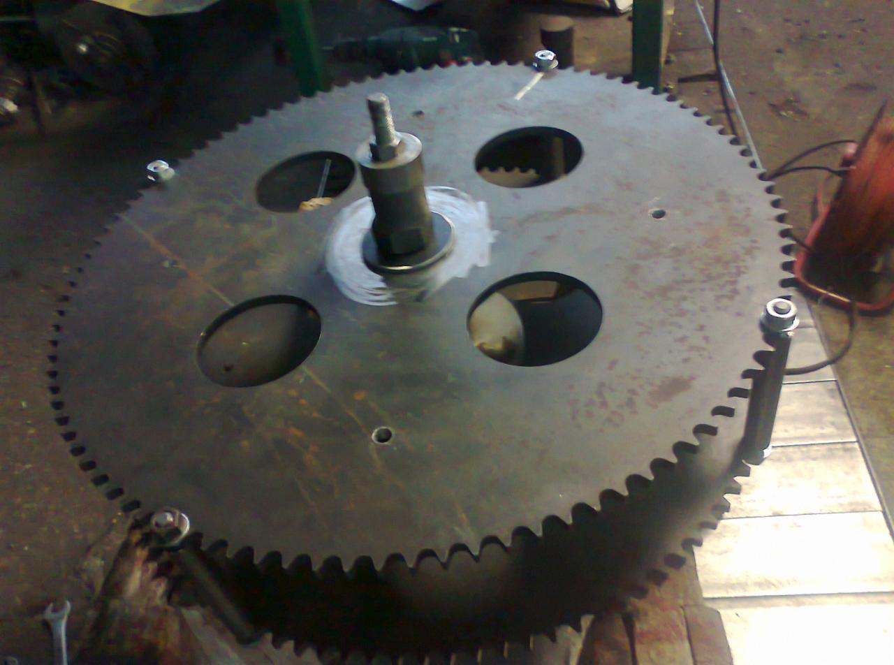 Duże, metalowe koło zębate z wyciętymi otworami, zamocowane na trzpieniu i przykręcone śrubami, prawdopodobnie w trakcie naprawy lub modyfikacji.