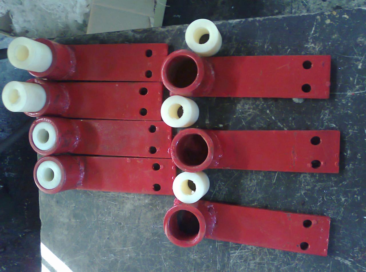 Czerwone, metalowe elementy z przyspawanymi uchwytami i włożonymi białymi tulejami z tworzywa sztucznego, ułożone rzędami na brudnym stole warsztatowym.