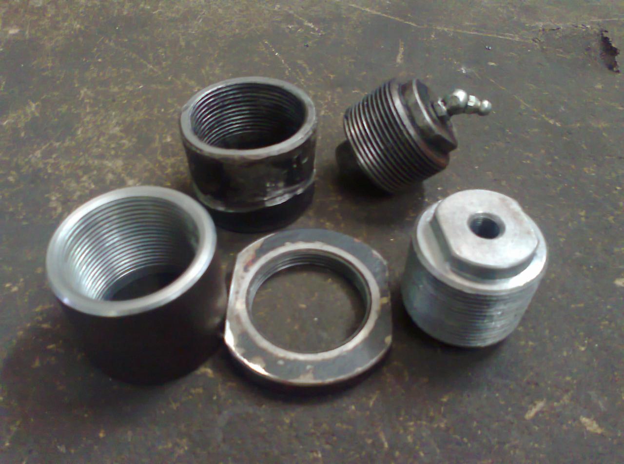 Zestaw metalowych elementów z gwintami, w tym tuleje, pierścień i element z kulką, leżących na szorstkiej, metalowej powierzchni.